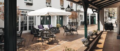 Un patio exterior con una zona de estar cubierta, una sombrilla blanca y plantas en macetas.