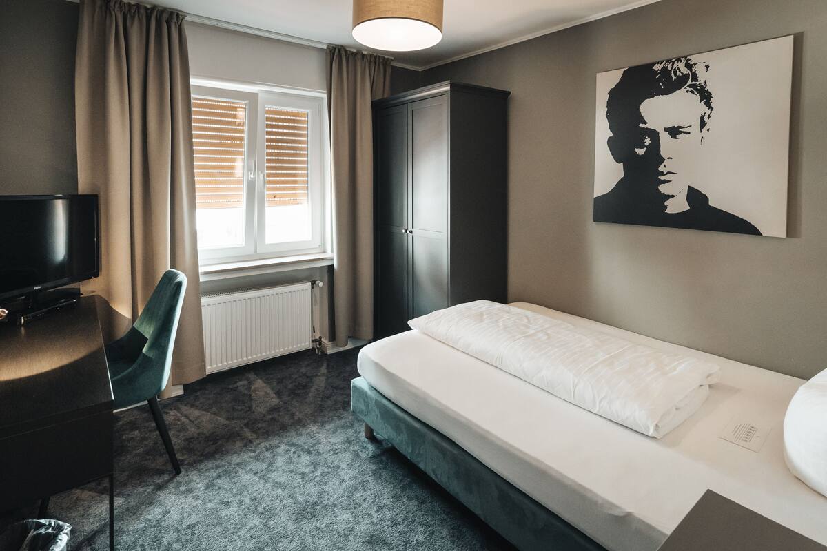 A hotel room with a bed, a desk, a chair, a wardrobe, and a window with blinds.