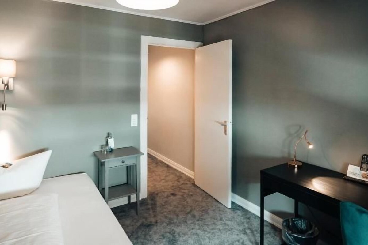 A hotel room with a bed, bedside table, lamp, and a door leading to another room.