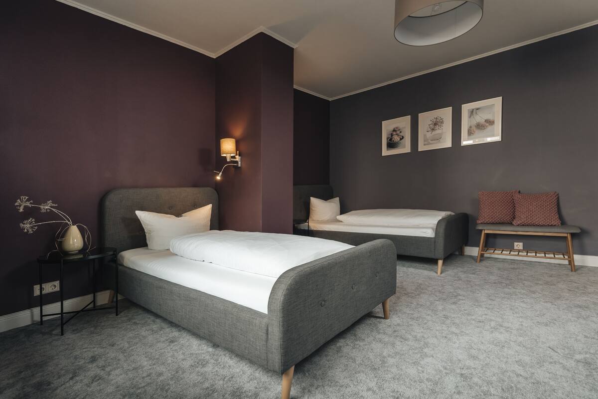 A modern bedroom with two beds, a bedside table, a wall-mounted lamp, and a bench.
