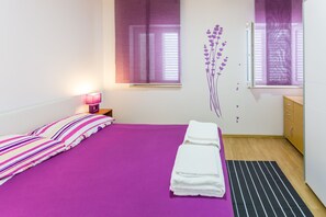 A bedroom with a purple bedspread, a bedside lamp, a window with blinds, a wooden dresser, and a wall decoration. - Apartments Dalmacija (Omis)