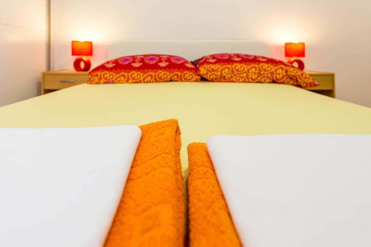 A neatly made bed with orange linens and two red patterned pillows.