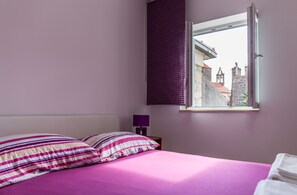 A bedroom with a bed, pillows, a nightstand, a lamp, and a window with a view of buildings. - Apartments Dalmacija (Omis)