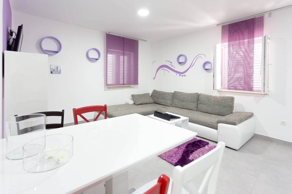A modern living room with a white sofa, grey cushions, a white coffee table, and a purple rug. There are two windows with purple blinds, a white ceiling, and a white wall with a purple decorative element.