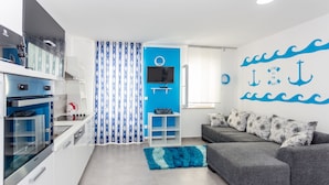 A modern kitchen with white cabinets and a built-in oven, a grey sofa with patterned cushions, and a blue accent wall with nautical decor. - Apartments Dalmacija (Omis)