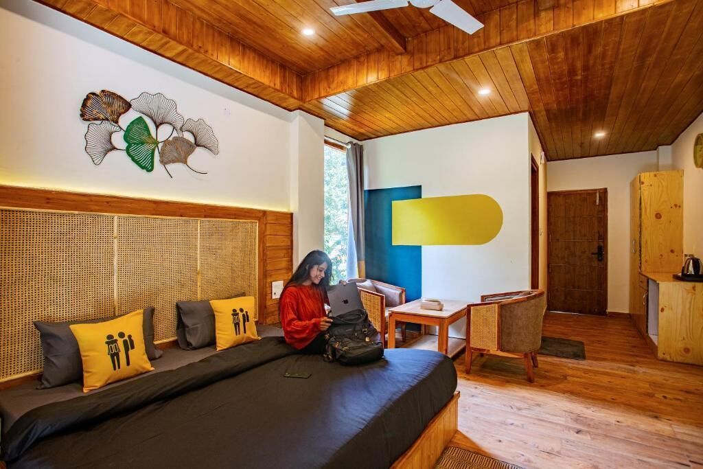 A room with a bed, a desk, and a chair. There is a person sitting on the bed.