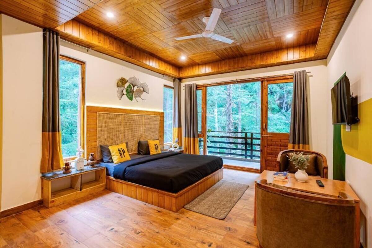 A bedroom with a wooden ceiling, a bed, a desk, and a large window with a view of trees.