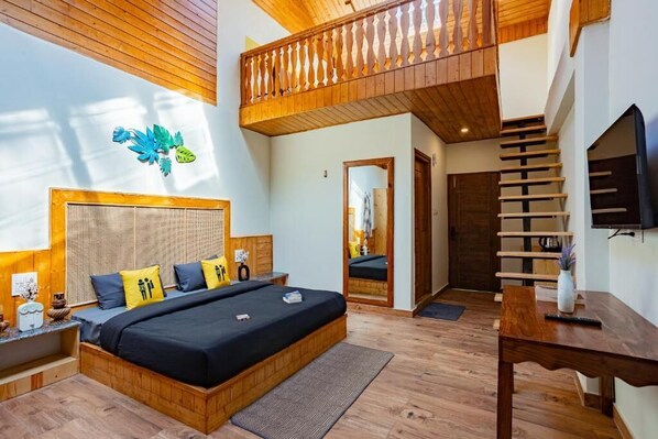 Un dormitorio con una cama grande, cabecero de madera, un balcón con barandilla de madera, un televisor y un espejo.
