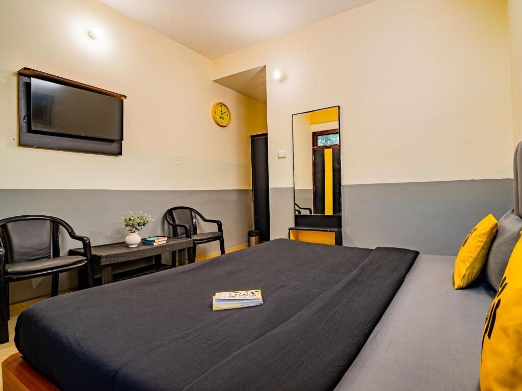 Hotellihuone, jossa on sänky, kaksi tuolia, pieni pöytä, jossa on maljakko ja kirjoja, seinälle asennettu televisio ja kello.
