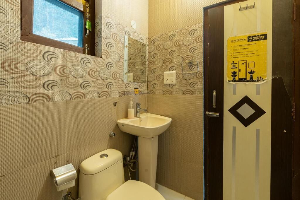 Casa de banho com sanita, lavatório e espelho. Há uma janela sobre o lavatório e uma porta com um aviso.
