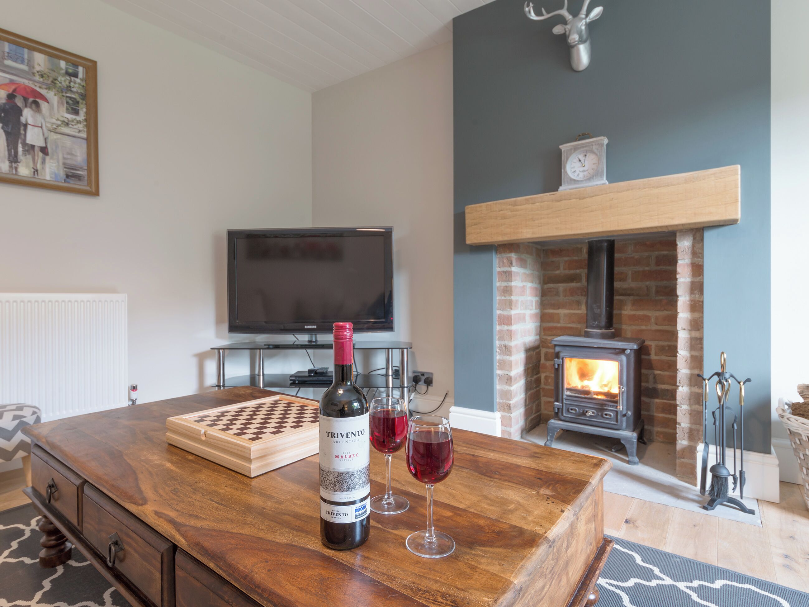Una sala de estar con una mesa de centro de madera, una botella de vino y una copa de vino tinto. Hay un televisor, una chimenea y un reloj en la pared.