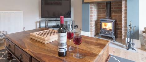 Una sala de estar con una mesa de centro de madera, una botella de vino y una copa de vino tinto. Hay un televisor, una chimenea y un reloj en la pared.