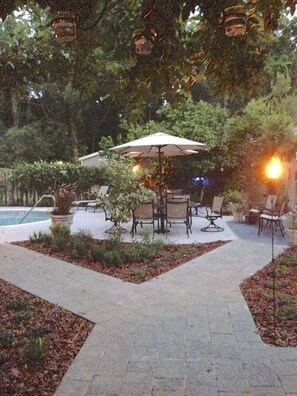 An outdoor patio area with a pool, surrounded by trees and plants. There are several tables and chairs under umbrellas. - Blue Heron Inn (Fernandina Beach)