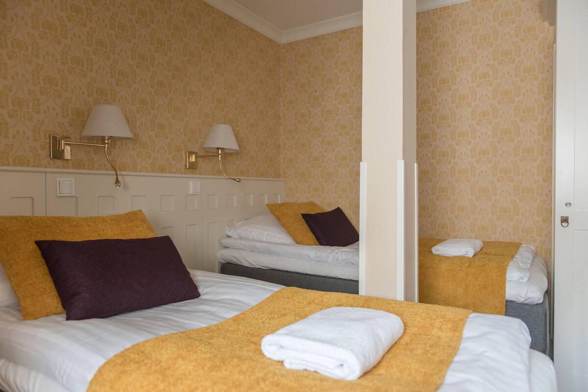 Habitación de hotel con dos camas, cada una con edredón blanco, manta marrón y almohada morada. Hay lámparas fijadas a la pared a ambos lados de las camas.