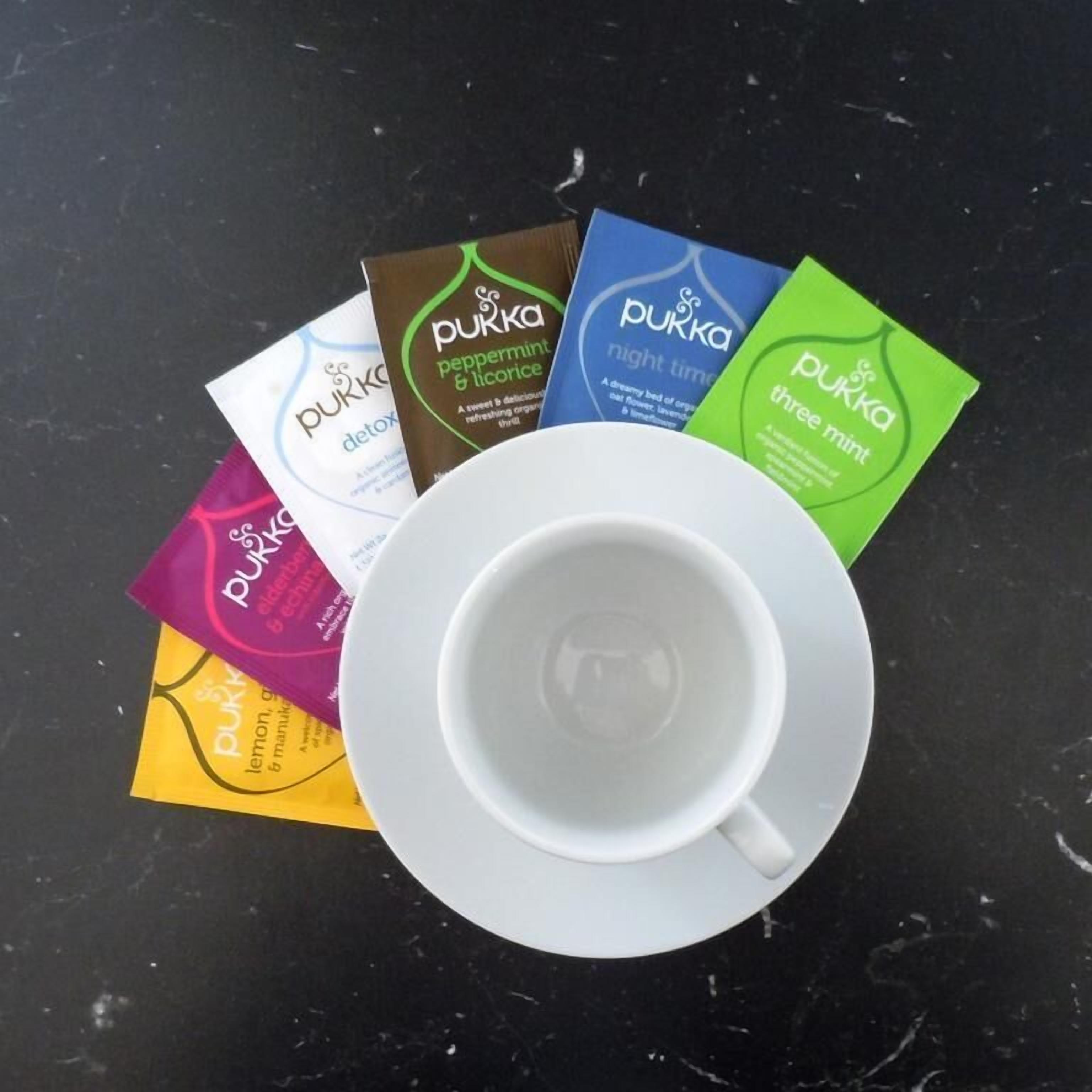 A white cup on a saucer with several Pukka tea bags in various flavors.