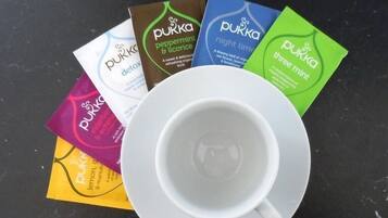 A white cup on a saucer with several Pukka tea bags in various flavors.