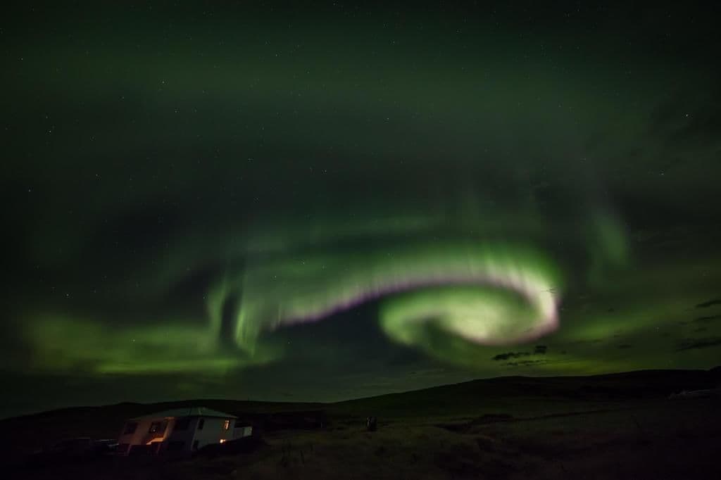 Una casa con porche visible y una estructura anexa, posiblemente un garaje o cobertizo, bajo un cielo nocturno con auroras boreales.