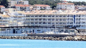 Un bâtiment situé sur la côte, doté de nombreux balcons, une enseigne indiquant « L’ESCALA BENVINGUTS BIENVENIDOS WELCOME », et une côte rocheuse au premier plan.