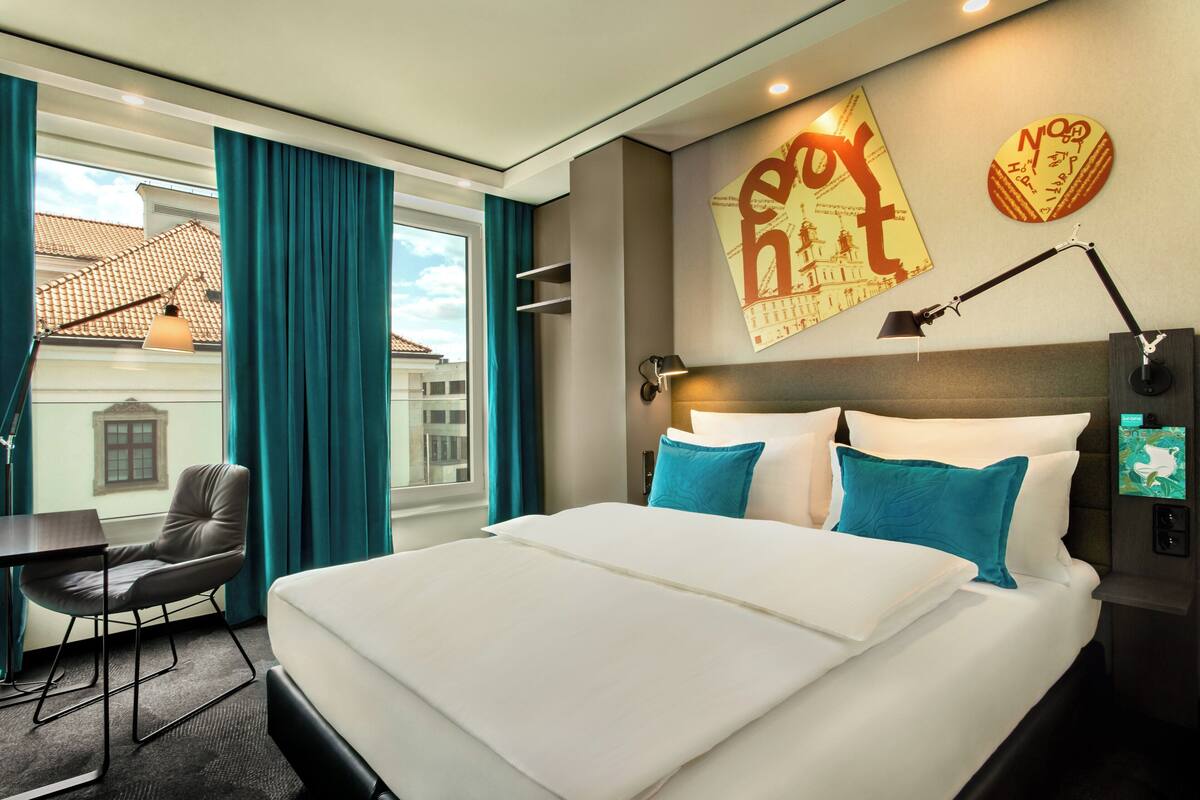 A hotel room with a large bed, a desk, and a chair. There is a window with curtains, a wall-mounted lamp, and a wall decoration.