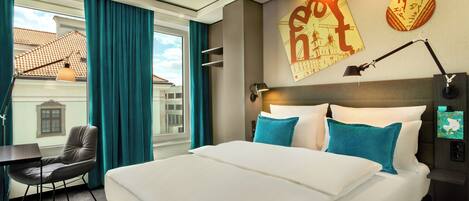 Habitación de hotel con una cama grande, un escritorio y una silla. Hay una ventana con cortinas, una lámpara de pared y un cartel en la pared.