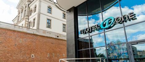A modern hotel entrance with a glass facade reflecting the sky and clouds.