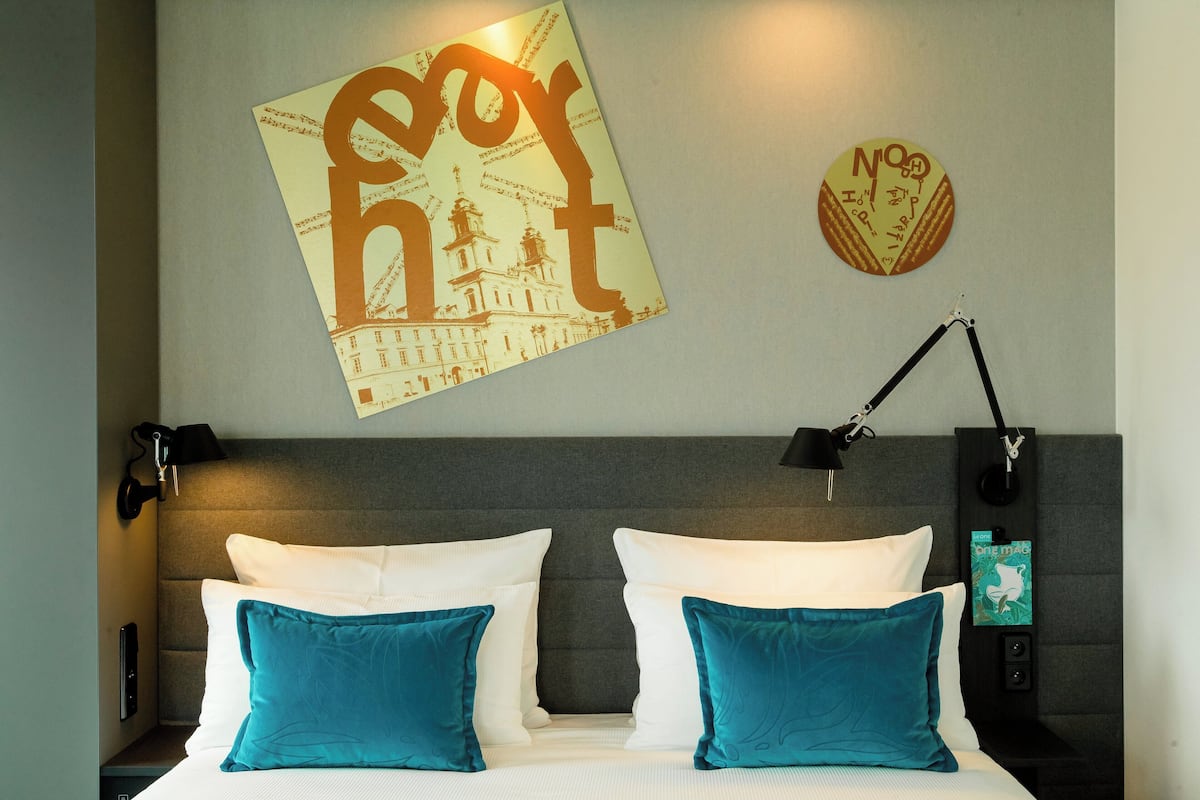 A hotel room with a bed, two teal pillows, a wall-mounted lamp, and a poster of a building.