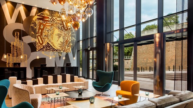 A modern lounge area with seating arrangements, a large illuminated wall art piece, and large windows offering a view of the outside. - Motel One Warsaw-Chopin (Warsaw)