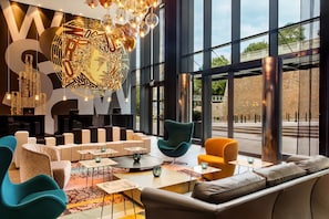 A modern lounge area with seating arrangements, a large illuminated wall art piece, and large windows offering a view of the outside. - Motel One Warsaw-Chopin (Warsaw)