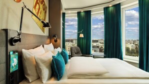A hotel room with a large bed, a desk, a chair, a city view, and a window with curtains. - Motel One Warsaw-Chopin (Warsaw)