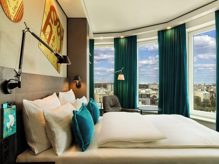 A hotel room with a large bed, a desk, a chair, a city view, and a window with curtains.