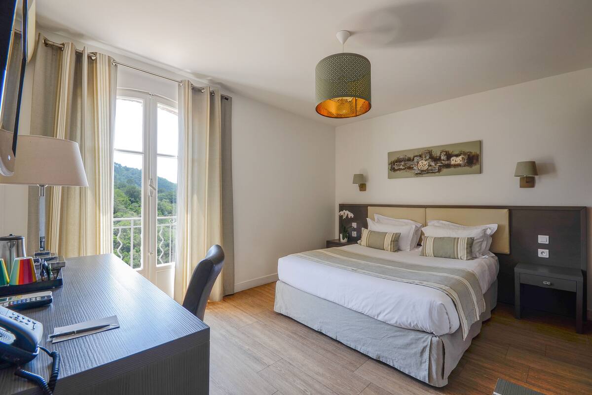A hotel room with a large bed, a desk with a telephone, and a view of greenery outside.