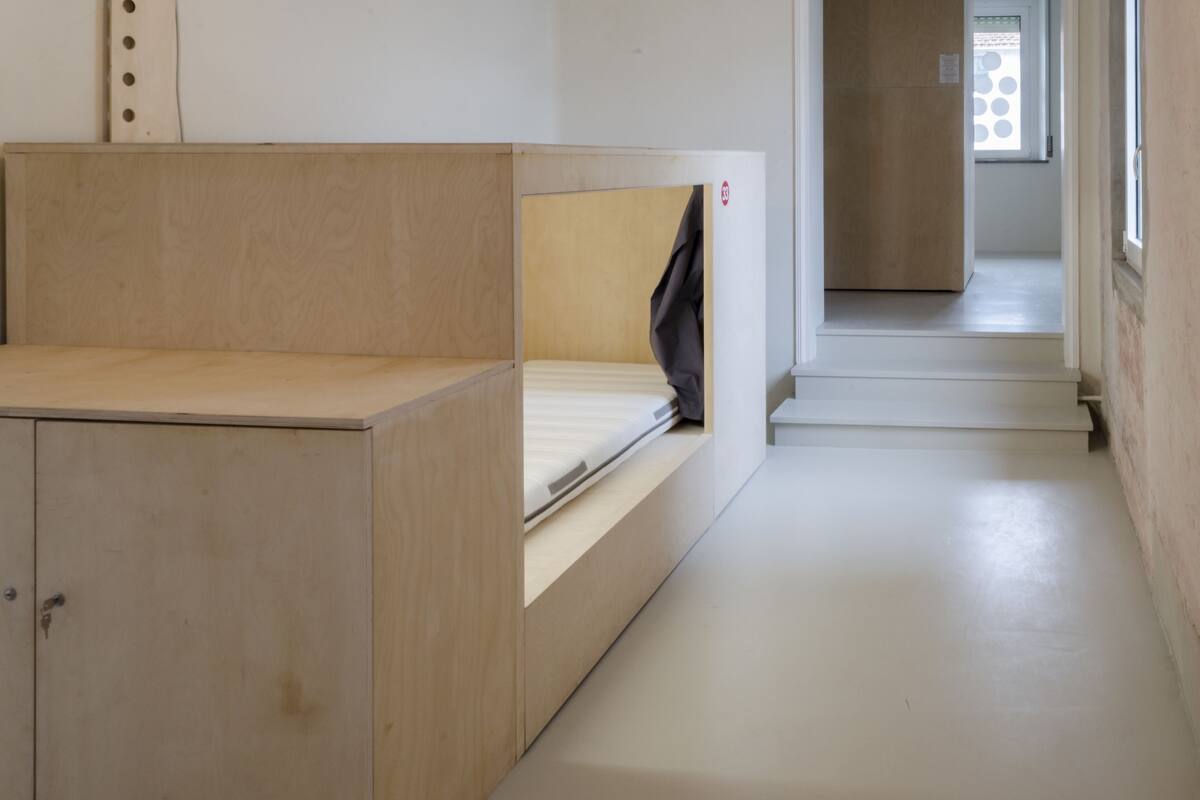A bunk bed with a wooden headboard and a single mattress. There is a door with a handle and a small window with circular patterns.