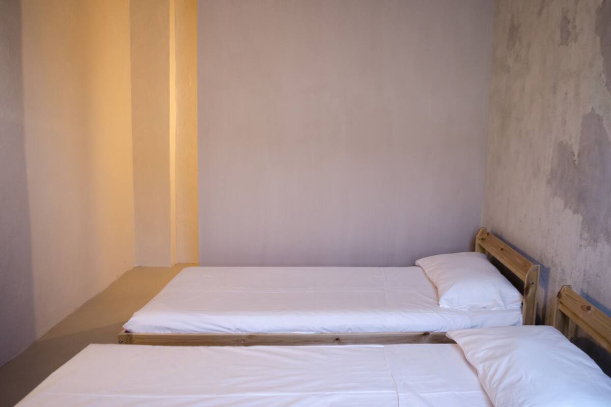 A small, dimly lit room with two single beds and a plain wall.