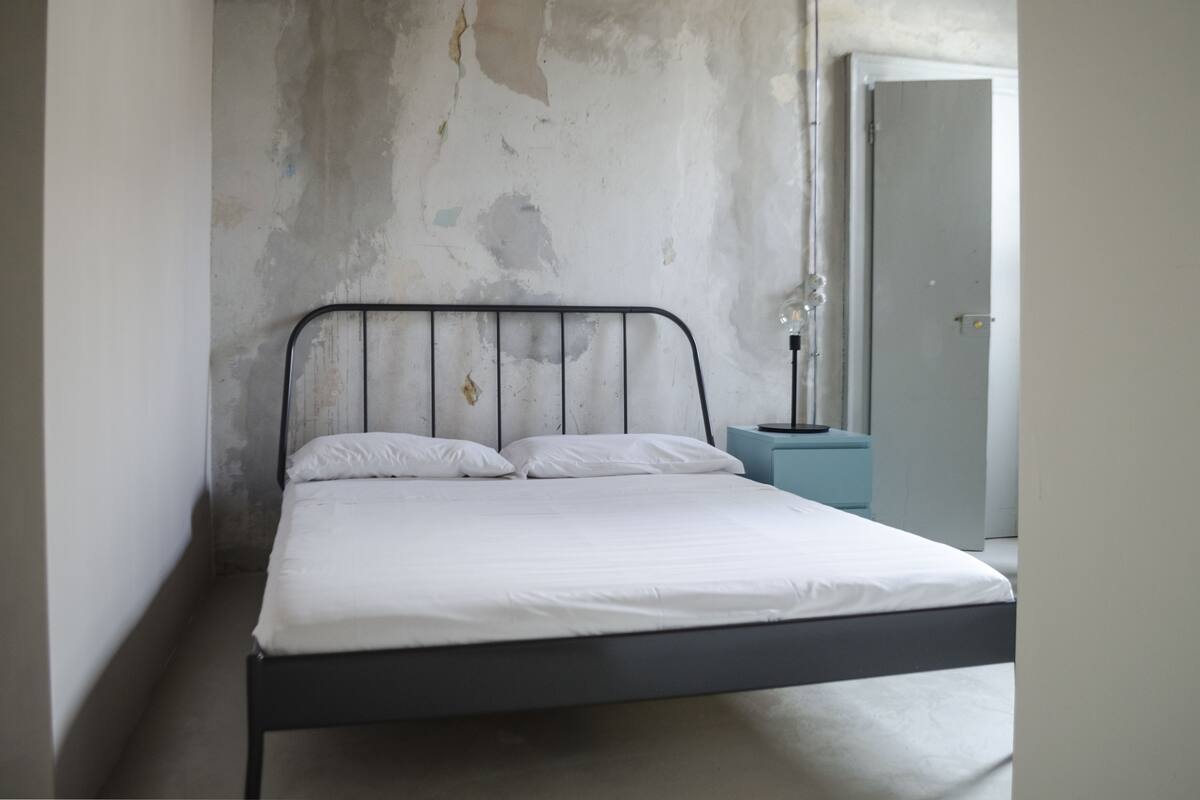 A single bed with a metal frame and white bedding in a room with a concrete wall and a door in the background.