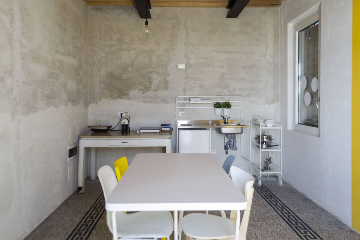 A compact kitchen with a white table, chairs, a small fridge, a sink, and a window with a view.