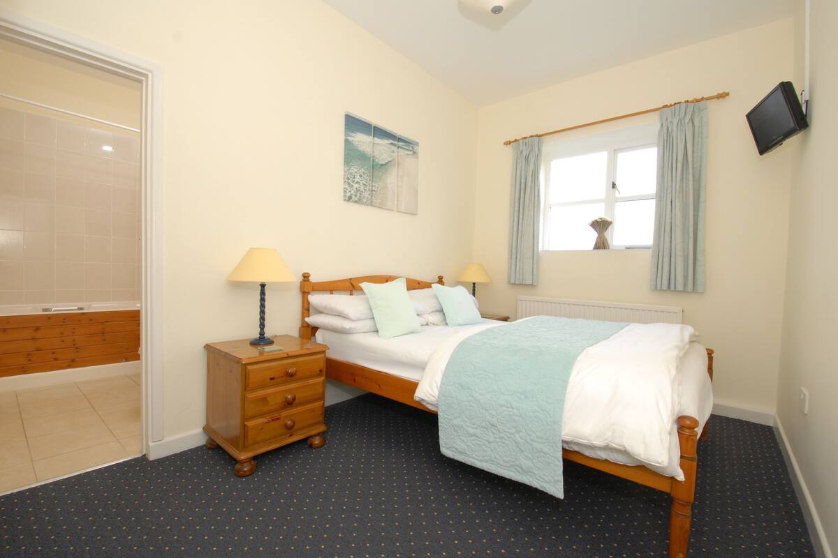 A bedroom with a bed, bedside lamps, a wooden nightstand, a window with curtains, and a wall-mounted TV.