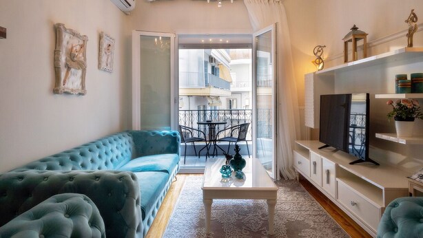 A living room with a blue sofa, a coffee table with decorative items, a TV on a white stand, and a balcony with a railing. - Acropolis Deluxe Apt Must (Thessaloniki)