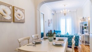 A dining area with a white table set for four, a chandelier, and a blue sofa in the background. - Acropolis Deluxe Apt Must (Thessaloniki)