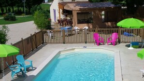 A rectangular swimming pool with clear blue water, surrounded by a wooden deck and a covered seating area with chairs and umbrellas.