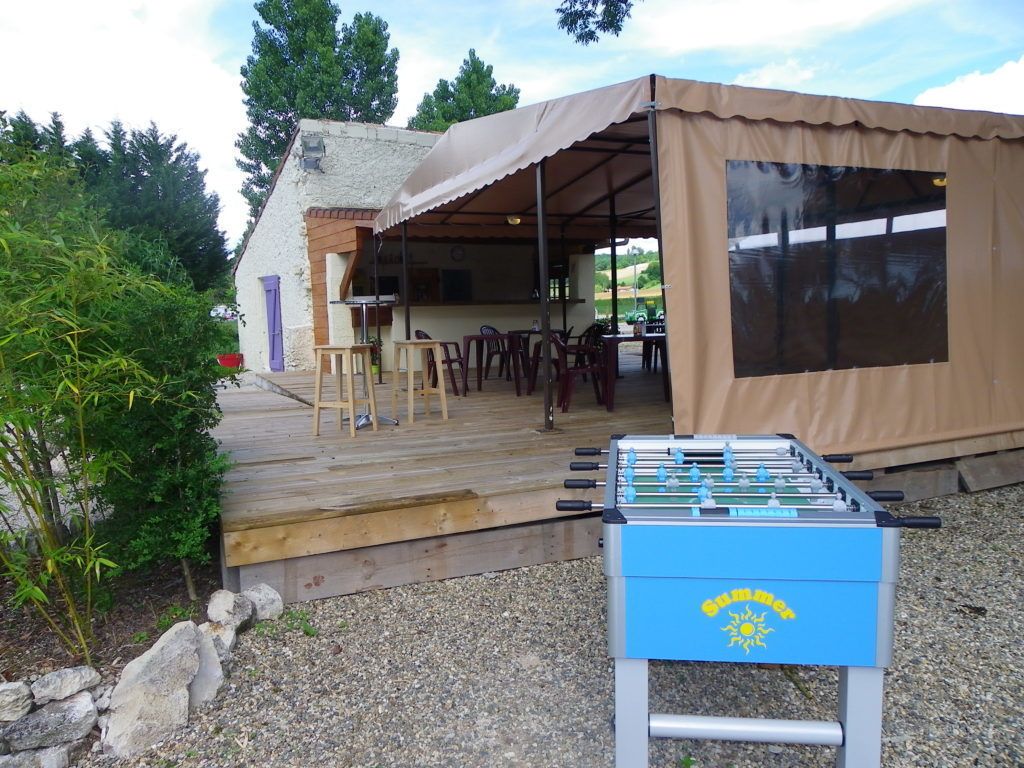 Une table de baby-foot est placée devant un espace extérieur abrité, aménagé avec des tables et des chaises.