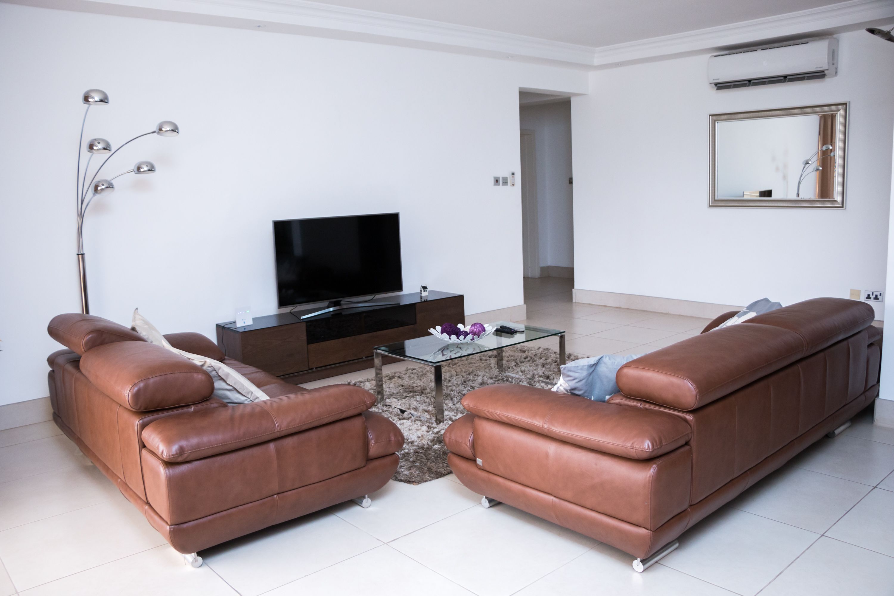 A modern living room with a brown leather sofa set, a flat-screen TV, a glass coffee table, and a floor lamp.