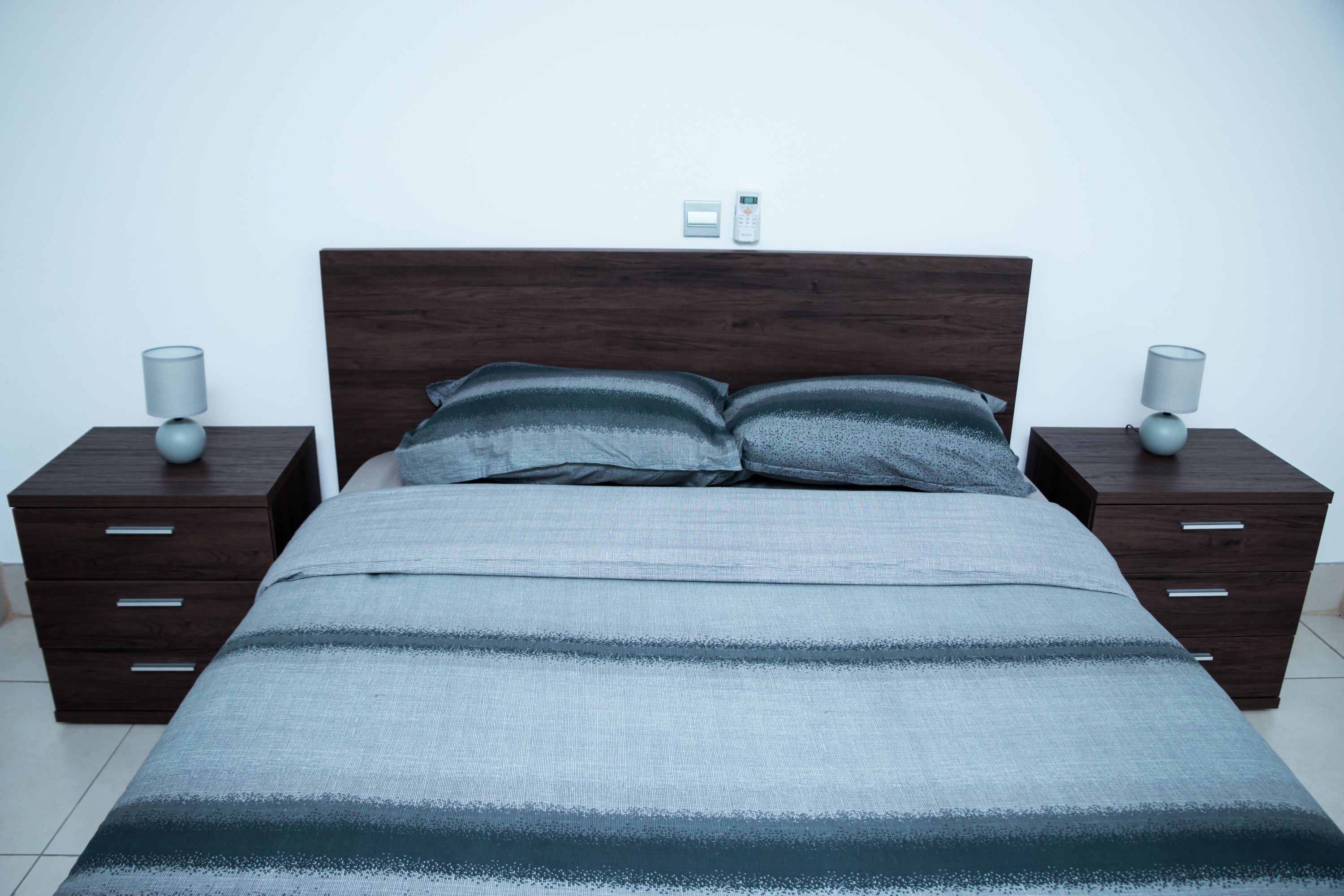 A neatly made bed with a striped bedspread, two bedside tables with lamps, and a wall-mounted thermostat.
