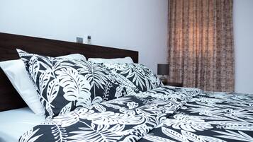 Una cama bien tendida con una colcha floral en blanco y negro, un cabecero de madera y una cortina marrón al fondo.
