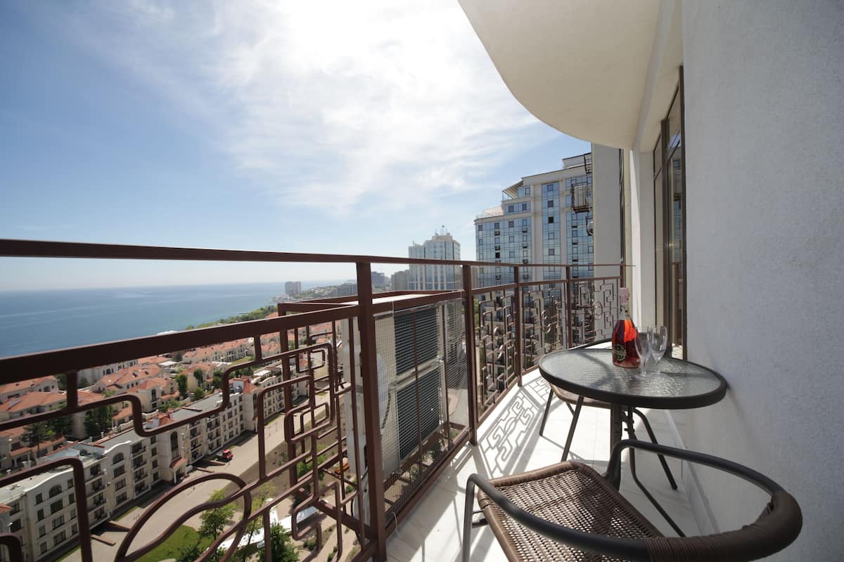 Uma varanda com vista para o mar e edifícios, uma mesa redonda com dois copos, e uma cadeira de vime.