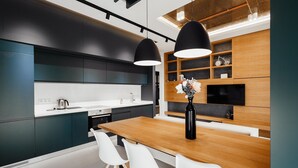A modern kitchen with dark cabinetry, a wooden dining table, white chairs, and pendant lighting. - Arcadia Dream Apartments (Odesa)