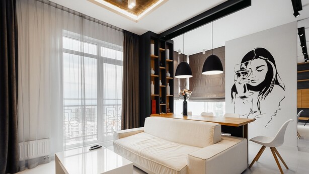 A modern living room with a sofa, a glass coffee table, and a dining area with a wooden table and chairs. There is a large window with sheer curtains, a bookshelf, and a wall-mounted artwork. - Arcadia Dream Apartments (Odesa)