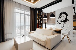 A modern living room with a sofa, a glass coffee table, and a dining area with a wooden table and chairs. There is a large window with sheer curtains, a bookshelf, and a wall-mounted artwork.