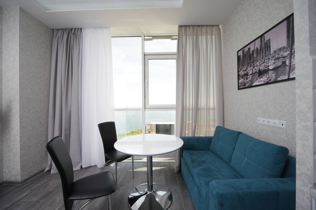 Quarto moderno com sofá verde-água, uma mesa redonda e duas cadeiras pretas. Há uma janela grande com cortinas, um quadro na parede e um aparelho de ar condicionado à vista.
