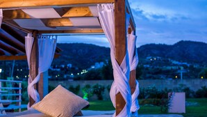 Uma cama de madeira com roupa de cama branca e um único travesseiro, com uma paisagem montanhosa ao fundo.