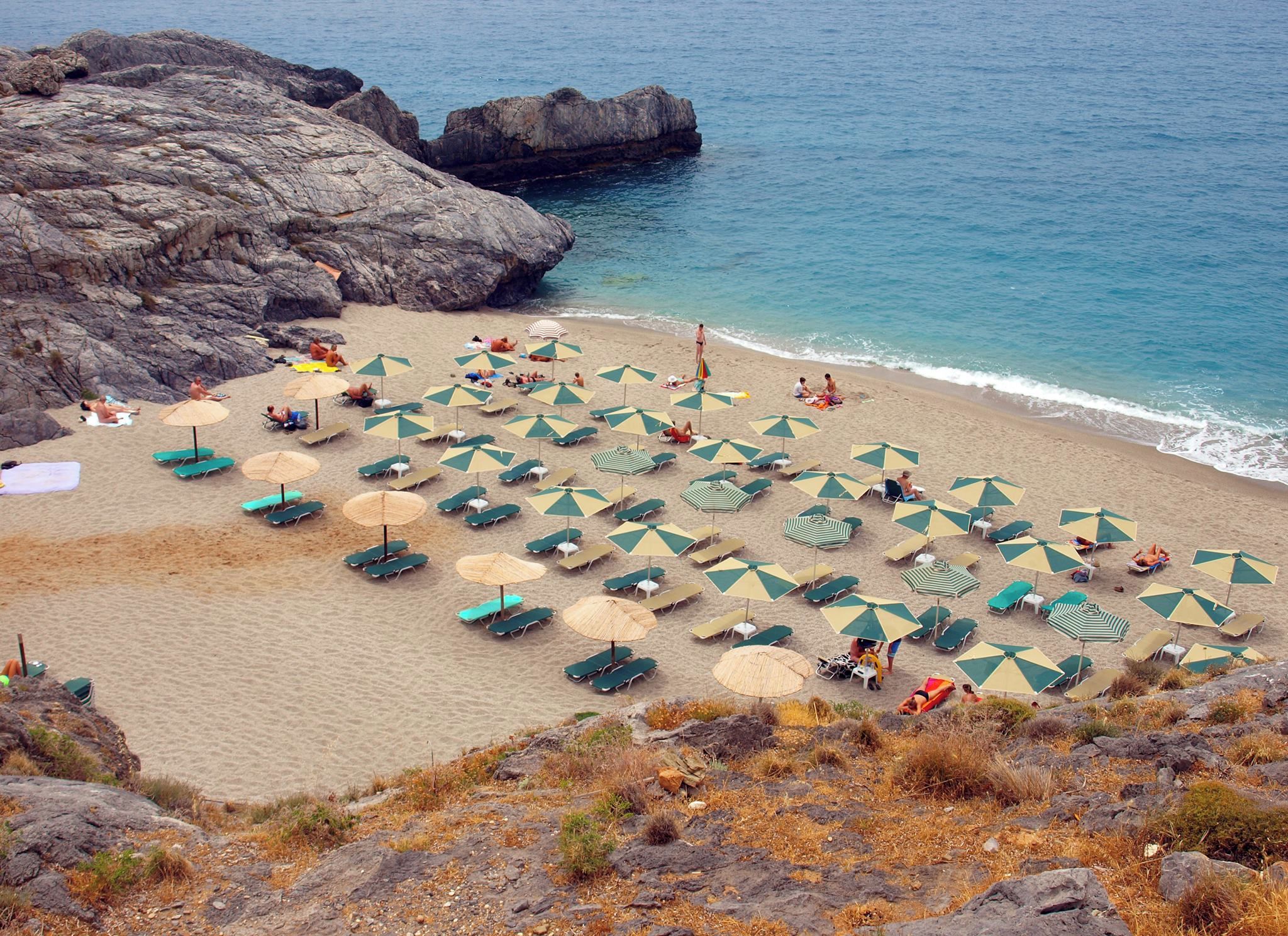 A beach with numerous umbrellas and lounge chairs, rocky terrain, and a view of the sea.